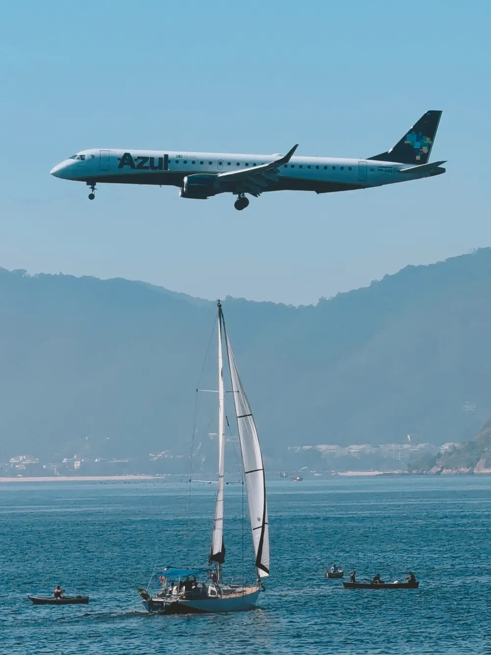Avião em aproximação sobre um veleiro e barcos a remo na baía