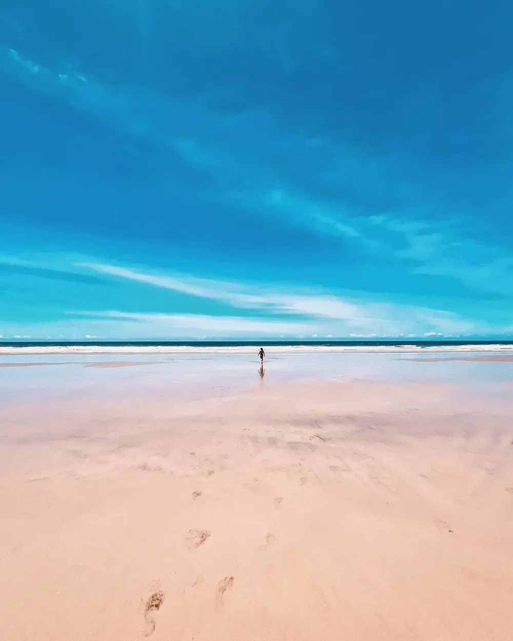 Pessoa sozinha caminhando numa praia imensa sob céu azul intenso