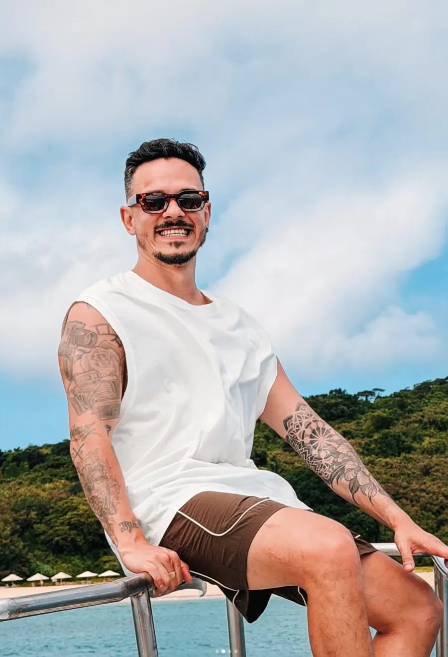 Marcos Jr. sorrindo de óculos escuros sentado na proa de um barco, com mata e mar ao fundo